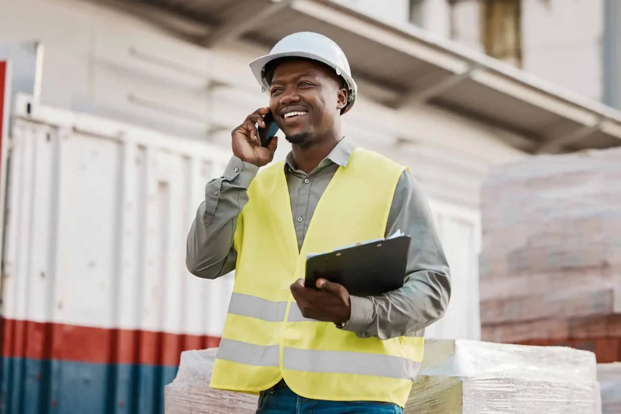 Worker in a yellow safety vest and helmet talks on the phone, holding a clipboard, at our Greenville location.