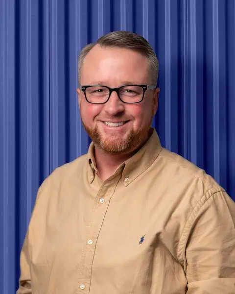 Smiling man in beige shirt against blue corrugated wall, associated with Carolina Chem Strip's industrial services.